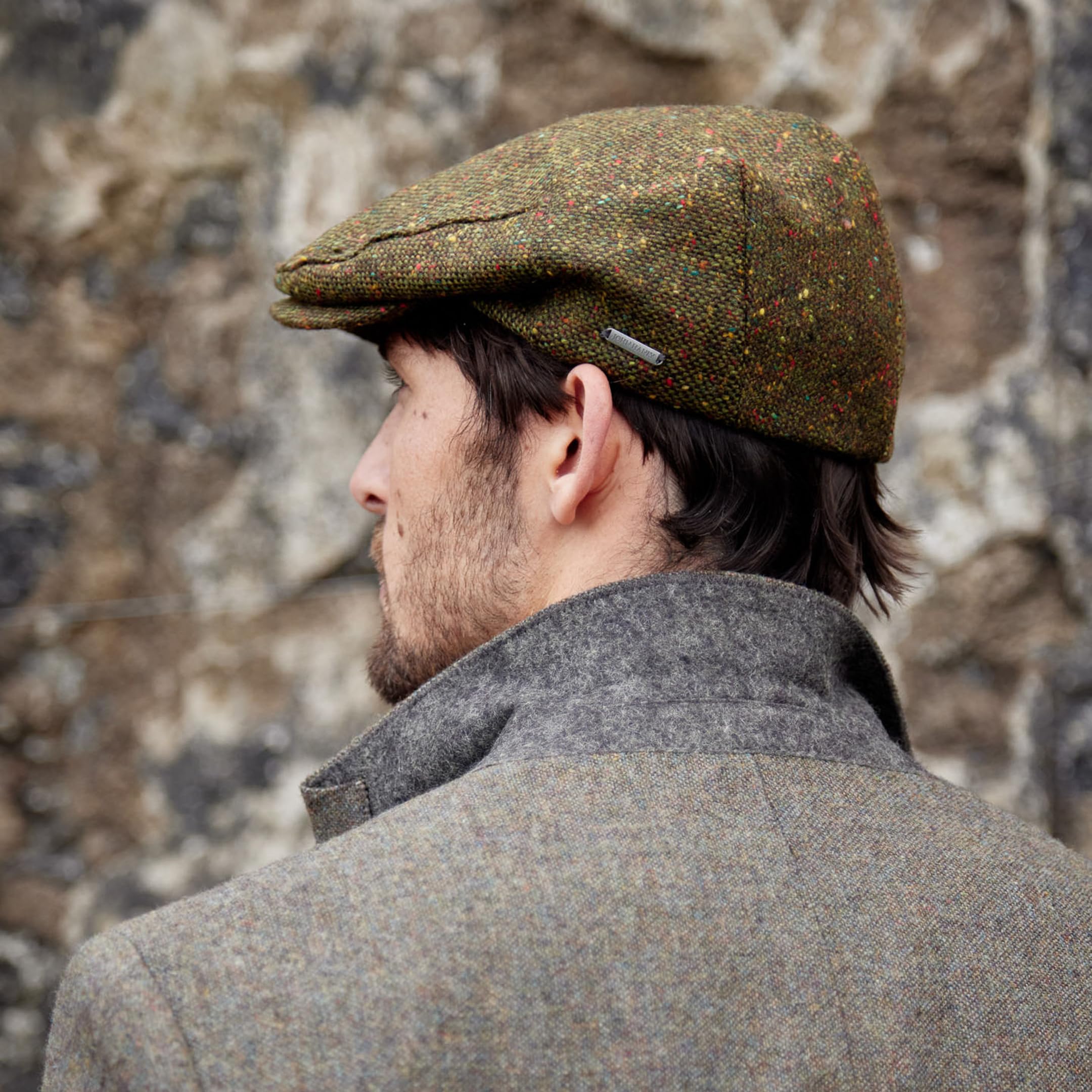 Man wearing a green tweed flat cap and gray coat against a stone wall.